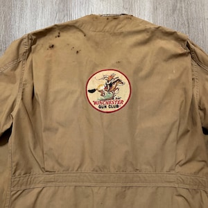 Peut inclure: Veste beige avec un écusson Winchester Gun Club. L'écusson représente un cavalier à cheval dans un motif circulaire avec des accents rouges et blancs. La veste a une fermeture boutonnée et un col.