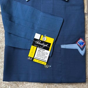 May include: A navy blue Pennleigh men's wear shirt with a light blue pocket and emblem. The shirt has a yellow tag with the brand name and details. The shirt is made of rayon and is washable.