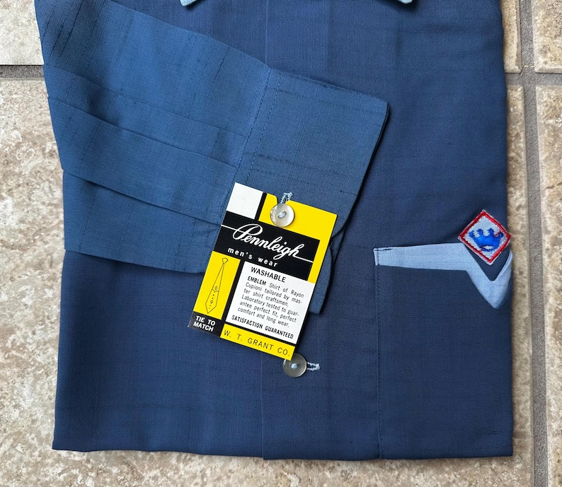 May include: A navy blue Pennleigh men's wear shirt with a light blue pocket and emblem. The shirt has a yellow tag with the brand name and details. The shirt is made of rayon and is washable.