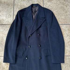 May include: A dark navy blue double-breasted wool coat featuring wide lapels, six black buttons arranged in two vertical rows, two front flap pockets, and shoulder epaulets. The interior is lined with a smooth, dark brown fabric.
