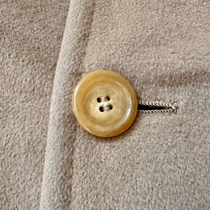 May include: Close-up of a tan coat with a large, round, light-colored button. The button is attached to the coat with a buttonhole. The coat fabric has a textured appearance.