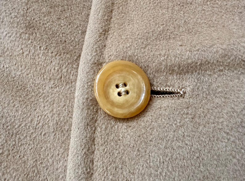 May include: Close-up of a tan coat with a large, round, light-colored button. The button is attached to the coat with a buttonhole. The coat fabric has a textured appearance.