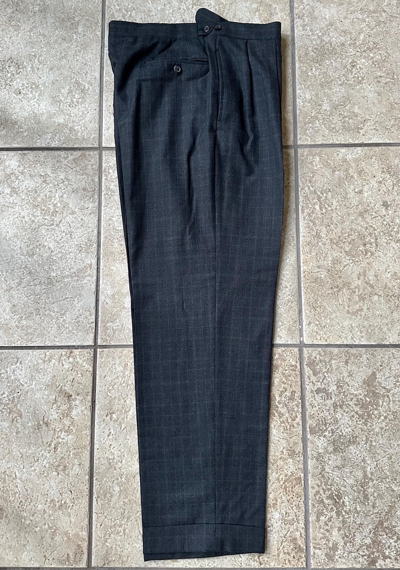 May include: A pair of dark gray dress pants with a subtle plaid pattern. The pants have a single pleat on each side and a button closure at the waist.