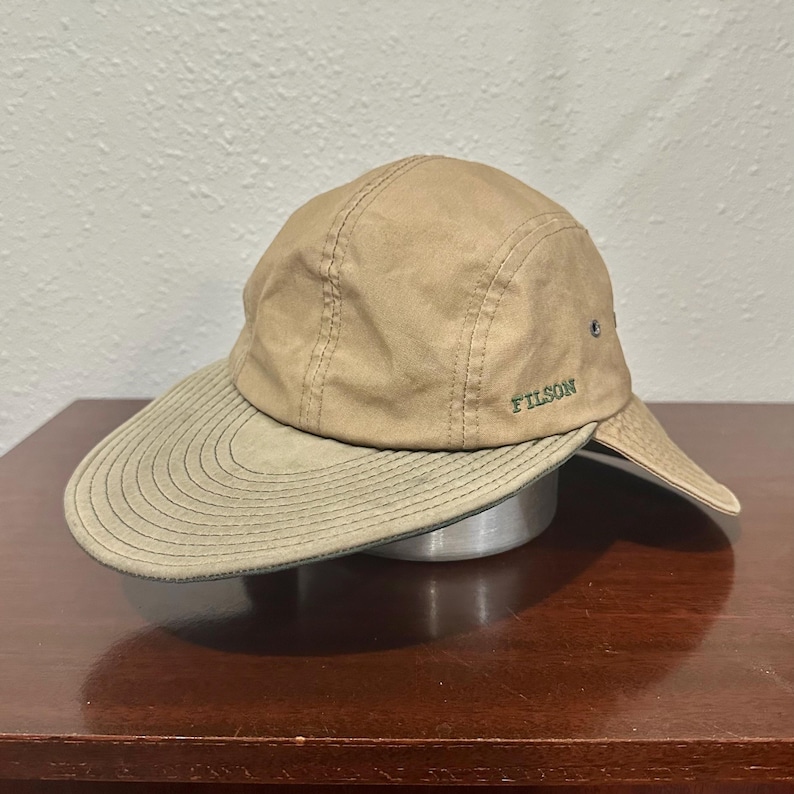 May include: A tan baseball cap with a wide, olive green brim featuring concentric stitching. The cap has a structured crown with visible seams and the word FILSON embroidered in green on the side. It includes a protective neck flap extension.