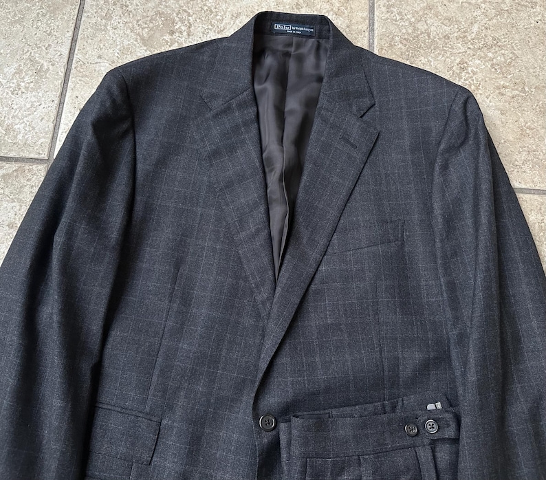 May include: A dark gray suit with a subtle plaid pattern. The suit jacket has a single button closure and a notched lapel. The suit pants have a flat front and a belt loop waistband.