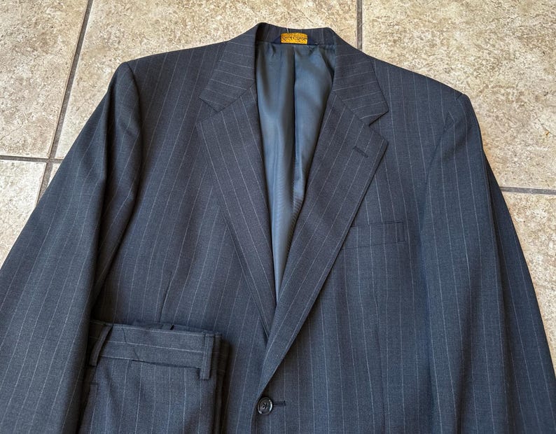 May include: A dark gray pinstripe suit jacket with a single button closure. The jacket has a notch lapel and a welt pocket on the left side.