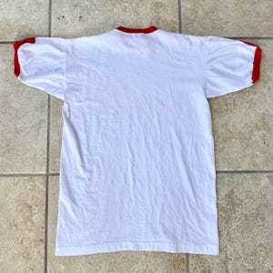 May include: A white t-shirt with red trim on the collar and sleeves. The shirt is made of a soft material and is laid flat on a tiled surface. The shirt is a classic style.