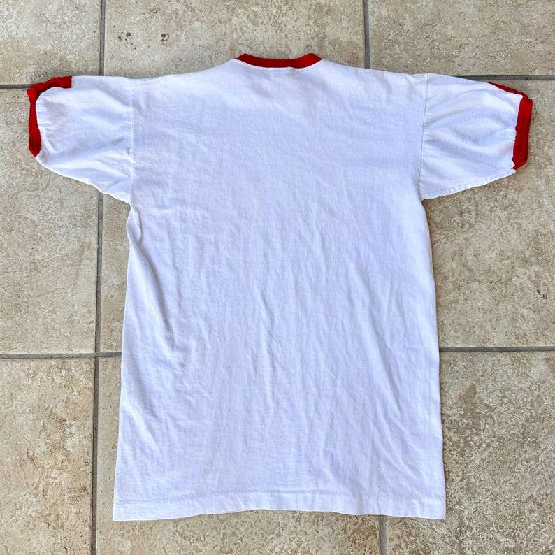 May include: A white t-shirt with red trim on the collar and sleeves. The shirt is made of a soft material and is laid flat on a tiled surface. The shirt is a classic style.