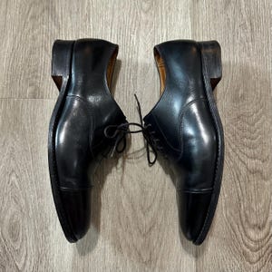 May include: A pair of black leather dress shoes with a cap toe design. The shoes have a classic Oxford style with laces and a polished finish. The heels are a dark brown colour.