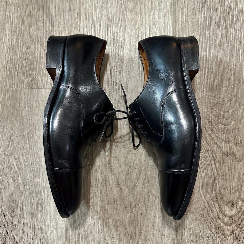 May include: A pair of black leather dress shoes with a cap toe design. The shoes have a classic Oxford style with laces and a polished finish. The heels are a dark brown colour.