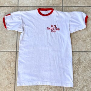 May include: White t-shirt with red trim on the collar and sleeves. The text "U.S. SOCCER CAMP STAFF" is printed in red on the front. The shirt has short sleeves and a crew neck.