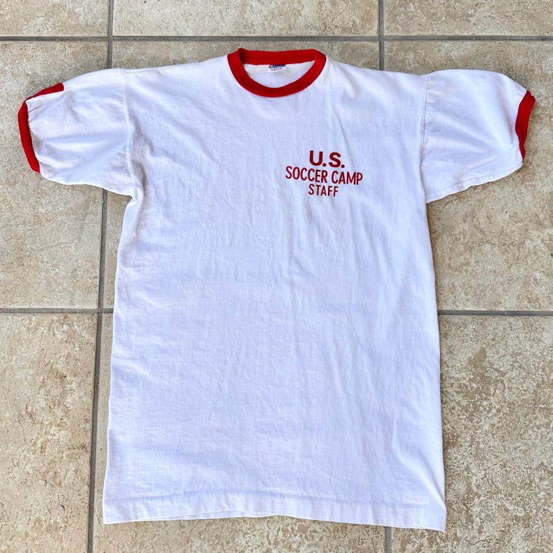 May include: White t-shirt with red trim on the collar and sleeves. The text "U.S. SOCCER CAMP STAFF" is printed in red on the front. The shirt has short sleeves and a crew neck.