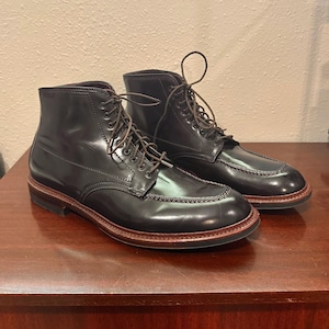 May include: A pair of dark brown leather boots with a polished finish, featuring a moc-toe construction, dark brown laces, and a stacked heel with a visible welt. The boots have a high-top silhouette with metal eyelets and a smooth, glossy surface.