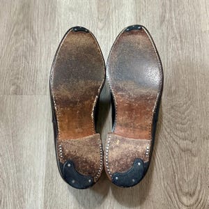 May include: A pair of well-worn, dark brown leather dress shoes, soles facing up. The soles show signs of wear and tear, with visible stitching and heel protectors. The shoes are set against a light wood grain background.
