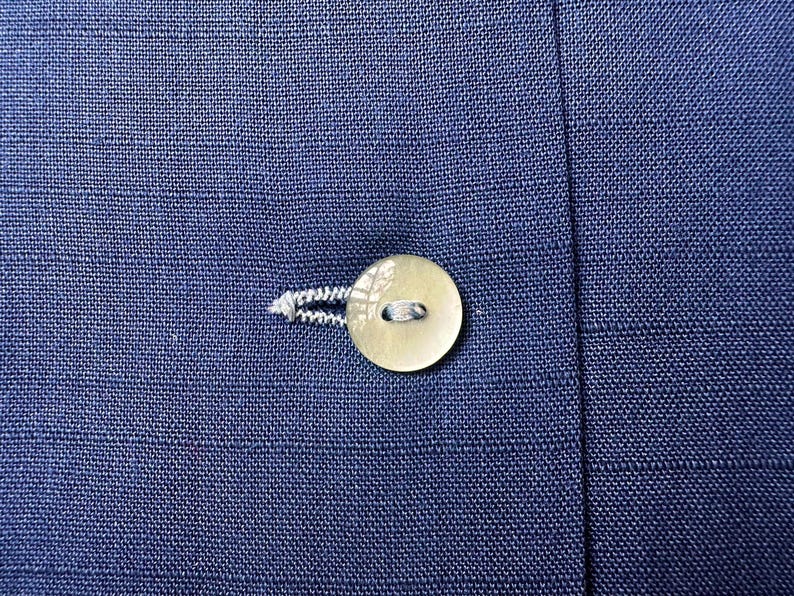 May include: Close-up of a navy blue fabric with a round, off-white button and a buttonhole. The button is attached with white thread. The fabric has a textured appearance.