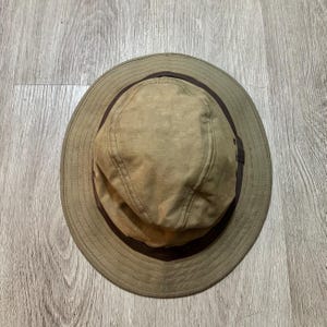 May include: A tan bucket hat featuring a wide brim and a dark brown band encircling the base of the crown. The hat is constructed with visible stitching along the brim and crown panels, and includes small metal ventilation eyelets on the sides.