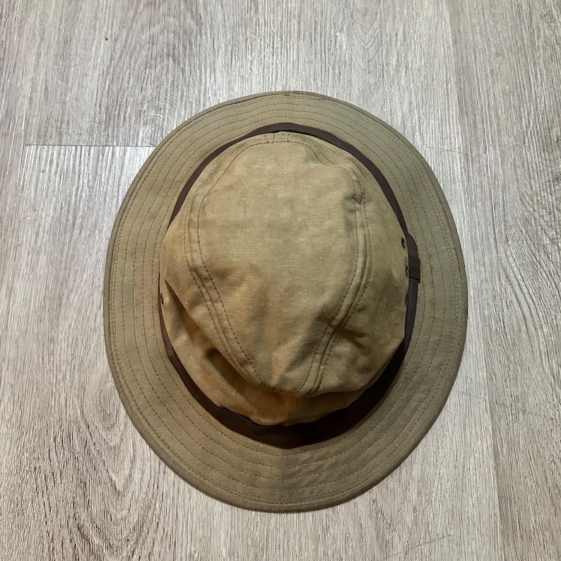 May include: A tan bucket hat featuring a wide brim and a dark brown band encircling the base of the crown. The hat is constructed with visible stitching along the brim and crown panels, and includes small metal ventilation eyelets on the sides.
