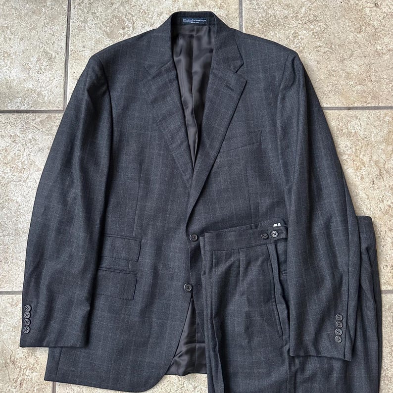 May include: A dark gray suit with a subtle plaid pattern. The jacket has a single button closure and the pants have a pleated front.
