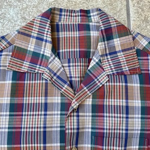 May include: A button-down shirt with a classic plaid pattern in shades of red, green, blue, and tan. The shirt features a collar and a chest pocket. The fabric appears to be a lightweight cotton or similar material.