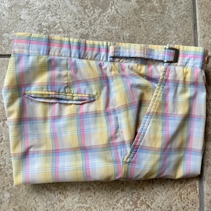 May include: Folded pair of shorts in a pastel plaid pattern. The shorts feature a yellow, pink, and blue plaid design with a buckle closure and a single back pocket. The fabric appears to be lightweight.