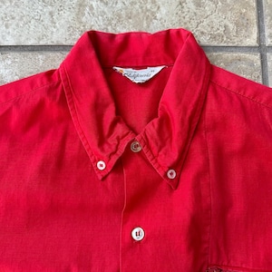 May include: A bright red button-down shirt with a classic collar and white buttons. The shirt has a small pocket detail on the lower right side. The tag reads "Made in California".