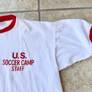 May include: White t-shirt with red trim on the collar and sleeves. The text "U.S. SOCCER CAMP STAFF" is printed in red on the front. The shirt is made of a soft, comfortable material.