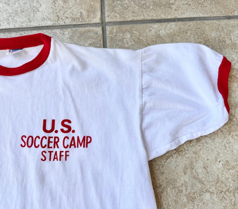 May include: White t-shirt with red trim on the collar and sleeves. The text "U.S. SOCCER CAMP STAFF" is printed in red on the front. The shirt is made of a soft, comfortable material.