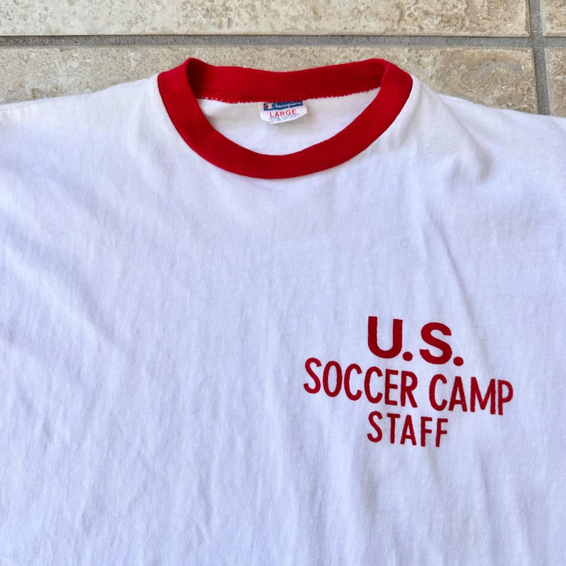May include: White t-shirt with a red collar and the text "U.S. SOCCER CAMP STAFF" in red. The shirt has a "LARGE" tag. The shirt is made of a soft material.