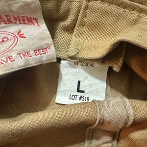 May include: A tan canvas garment featuring a white interior label with red text reading Filson Garment, Filson Co. Seattle, and You Don't Have To Hunt And Fish To Like Filson. A second white tag displays IN U.S.A., the letter L, and LOT #319.