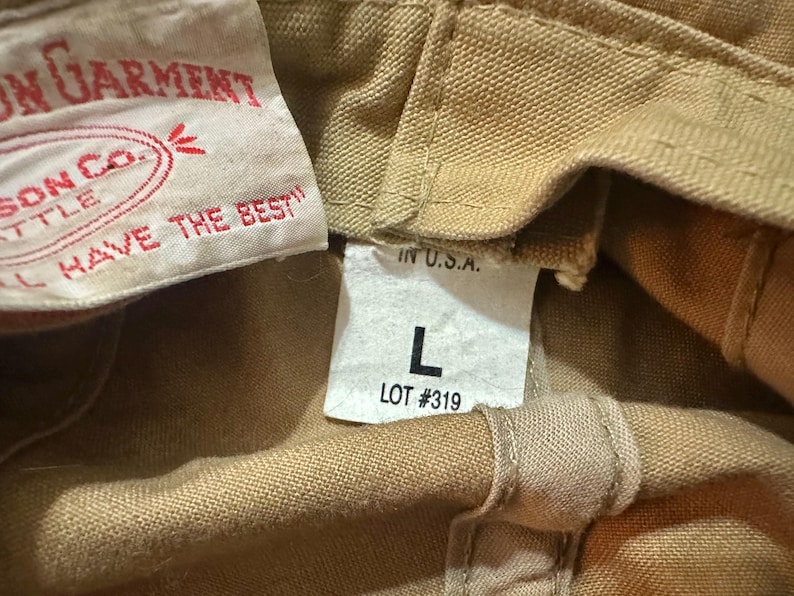 May include: A tan canvas garment featuring a white interior label with red text reading Filson Garment, Filson Co. Seattle, and You Don't Have To Hunt And Fish To Like Filson. A second white tag displays IN U.S.A., the letter L, and LOT #319.