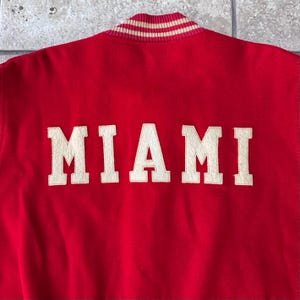 May include: Red varsity jacket with the word "MIAMI" in large, off-white letters across the back. The collar is striped with cream and tan. A classic, vintage style.