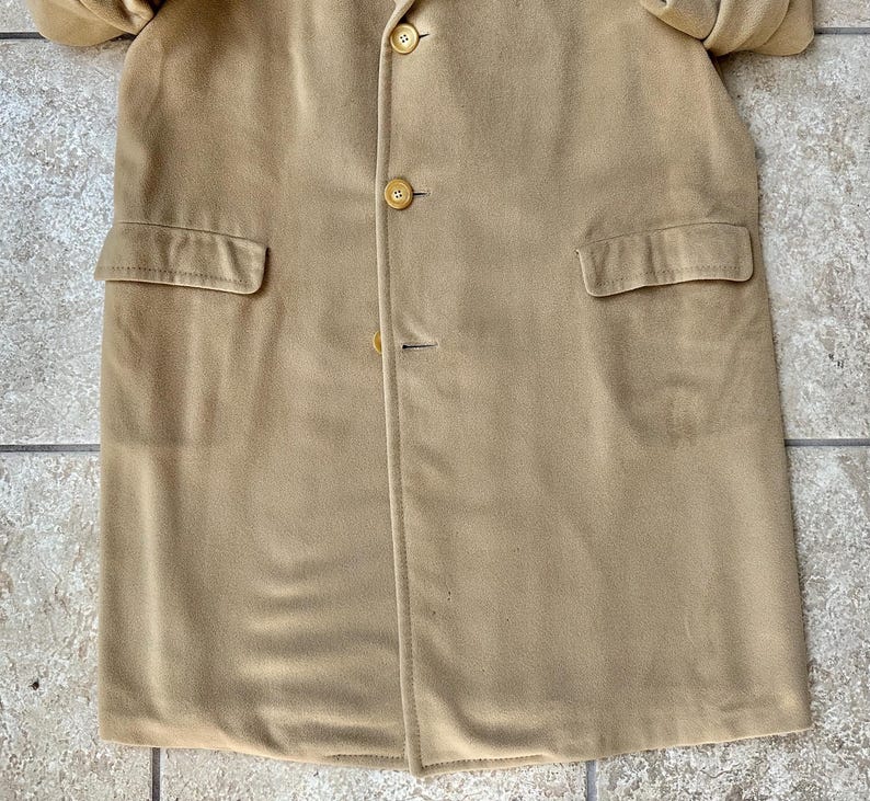 May include: A tan-colored coat with three buttons and two front pockets. The coat is made of a soft, suede-like material. The coat is buttoned up and appears to be in good condition.