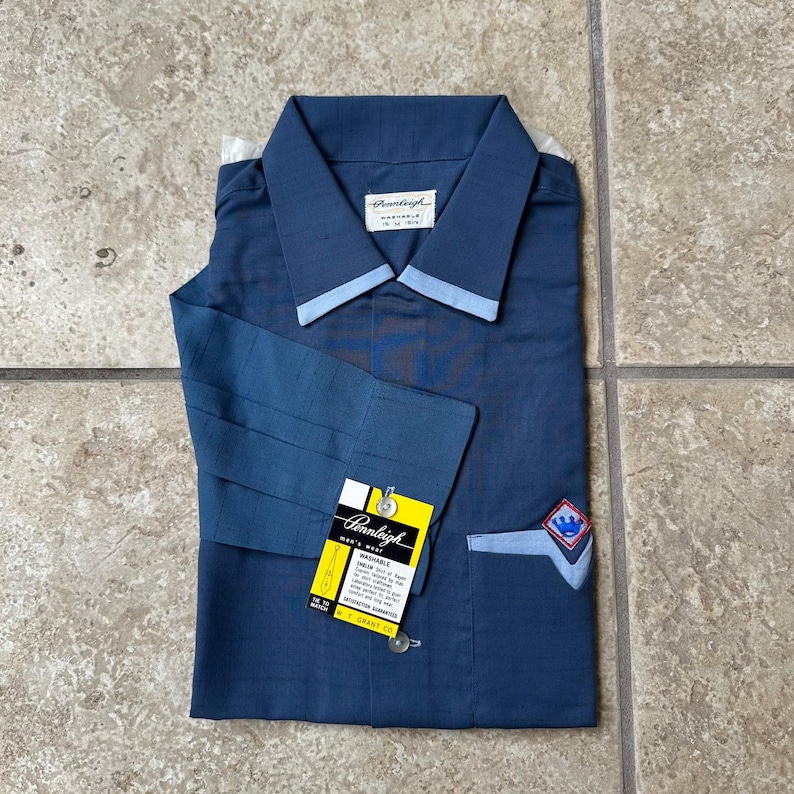 May include: A blue button-down shirt with a light blue collar and cuffs. The shirt has a pocket with a decorative emblem and a yellow tag with the brand name "Pennleigh". The shirt is folded neatly.