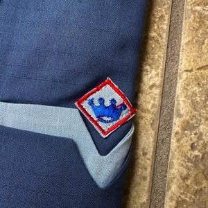 May include: Close-up of a navy blue fabric with a light blue accent and a diamond-shaped patch. The patch has a red border and features a blue crown design. The fabric appears to be part of a garment.