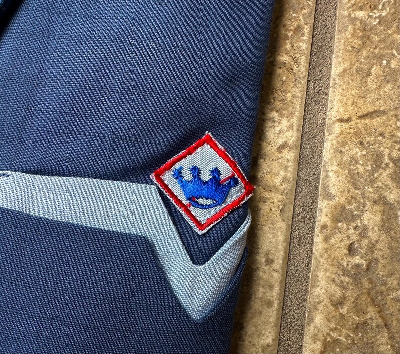 May include: Close-up of a navy blue fabric with a light blue accent and a diamond-shaped patch. The patch has a red border and features a blue crown design. The fabric appears to be part of a garment.