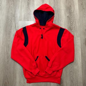 May include: A red hooded sweatshirt with a navy blue interior and accents. The sweatshirt features a front pocket, a button-up neckline, and navy blue stripes on the sleeves. The garment is made of a soft material.
