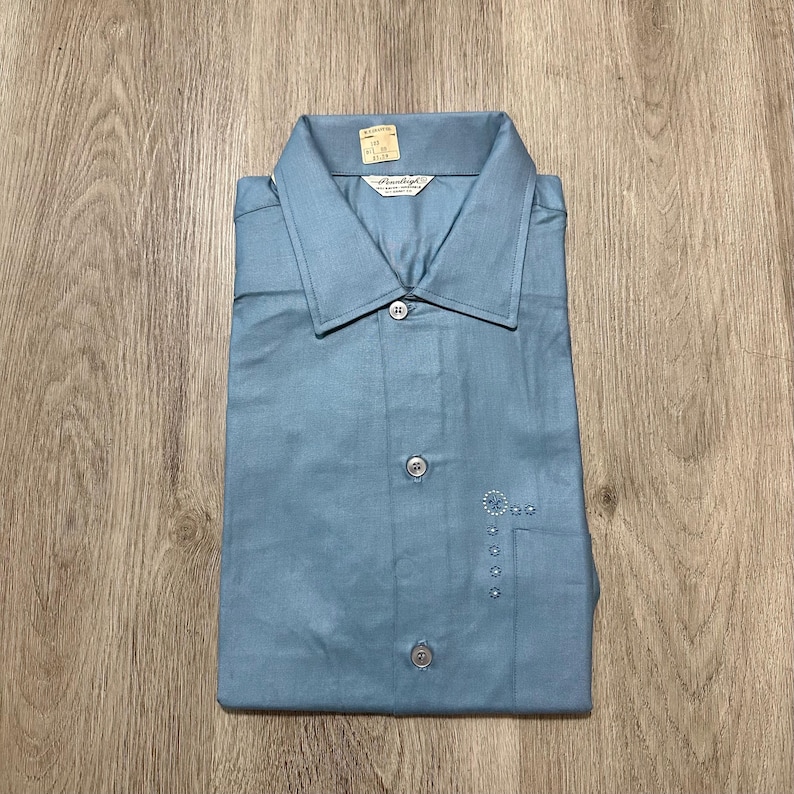 May include: A light blue button-down shirt with a pointed collar and a front placket featuring three visible buttons. The shirt includes a small embroidered floral design on the left chest area and a brand label inside the collar reading Pearly.