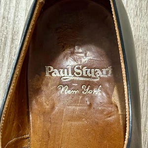 May include: Close-up of the interior of a black leather shoe. The shoe's insole is brown with the embossed text "Paul Stuart New York" in a silver colour. The shoe's leather is worn.