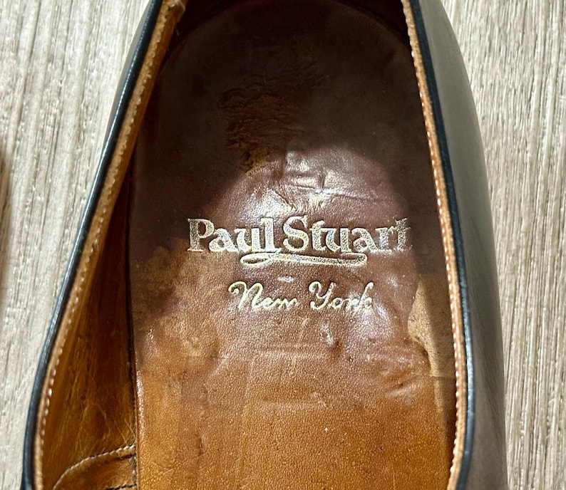 May include: Close-up of the interior of a black leather shoe. The shoe's insole is brown with the embossed text "Paul Stuart New York" in a silver colour. The shoe's leather is worn.