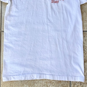 May include: A white t-shirt with the word "STAFF" printed in red at the top. The shirt is made of a soft, lightweight fabric and has a simple, classic design. The shirt is folded and lying on a light brown surface.