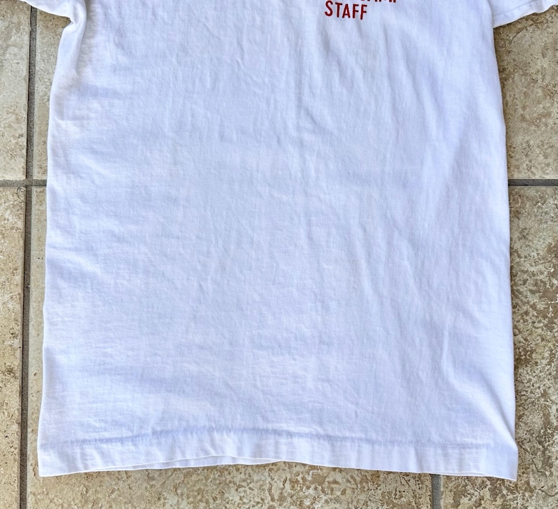 May include: A white t-shirt with the word "STAFF" printed in red at the top. The shirt is made of a soft, lightweight fabric and has a simple, classic design. The shirt is folded and lying on a light brown surface.