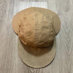 May include: A tan five-panel cap featuring a wide, circular brim that extends around the sides and back. The crown is constructed from multiple fabric panels with visible stitching, and two small metal ventilation eyelets are on the side.