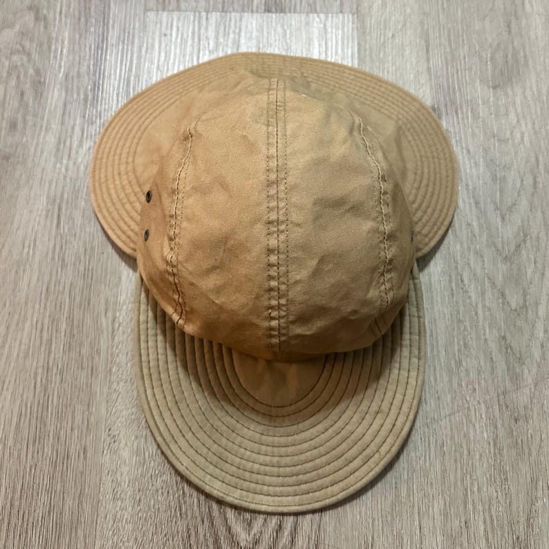 May include: A tan five-panel cap featuring a wide, circular brim that extends around the sides and back. The crown is constructed from multiple fabric panels with visible stitching, and two small metal ventilation eyelets are on the side.