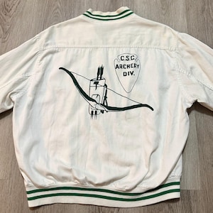 May include: A white varsity jacket with green striped ribbed trim at the collar and hem. The back features a dark green graphic of a bow and quiver with arrows, alongside a shield-shaped emblem containing the text C.S.C. ARCHERY DIV.