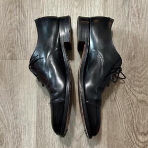 May include: A pair of black leather Oxford shoes with cap toes, laces, and stacked heels. The shoes are shown side-by-side, highlighting their polished finish and traditional style. The shoes are on a wooden surface.