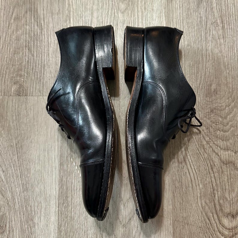 May include: A pair of black leather Oxford shoes with cap toes, laces, and stacked heels. The shoes are shown side-by-side, highlighting their polished finish and traditional style. The shoes are on a wooden surface.