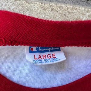 May include: Close-up of a white t-shirt with red trim around the neck and sleeves. A Champion brand tag is visible, with the word "LARGE" printed in red. The tag also states "100% COTTON".