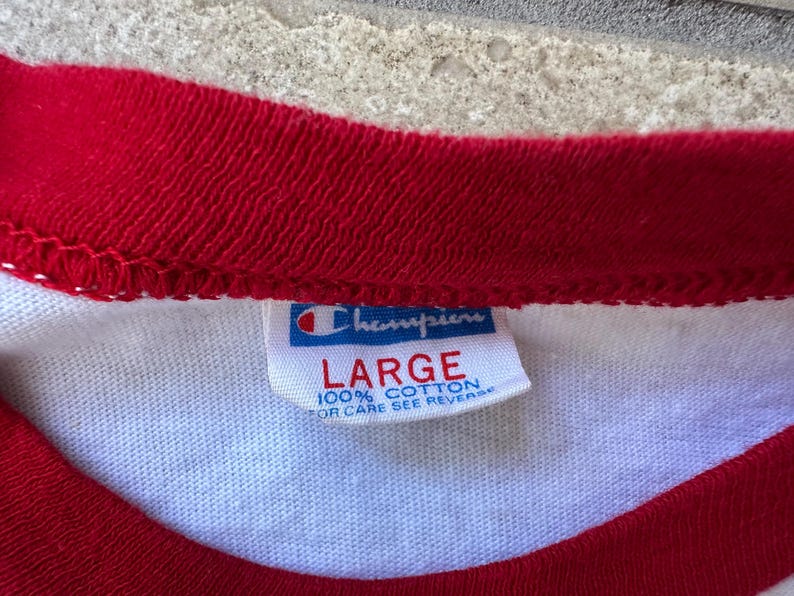 May include: Close-up of a white t-shirt with red trim around the neck and sleeves. A Champion brand tag is visible, with the word "LARGE" printed in red. The tag also states "100% COTTON".