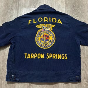 May include: A dark blue corduroy jacket featuring yellow embroidered text that reads FLORIDA and TARPON SPRINGS. Centered on the back is a large, circular yellow and blue emblem with an eagle, an owl, and the text VOCATIONAL AGRICULTURE FFA.
