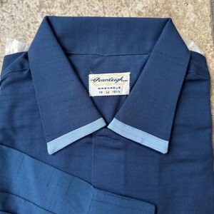 May include: A navy blue collared shirt with light blue trim on the collar and cuffs. The shirt has a label that reads "Pennleigh Washable 15 M 15 1/2". The shirt is folded neatly.